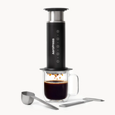 A black AeroPress steel with coffee dripping into a clear mug on a white background