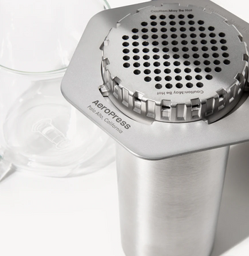 An AeroPress steel coffee maker with a stainless steel filter cap on a white background